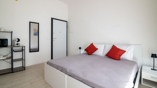 Um quarto moderno com cama, mesa de cabeceira, prateleira com cafeteira e uma porta.