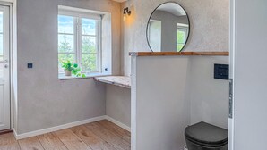 A bathroom with a wooden floor, a window with a plant, a round mirror, and a toilet. - Cozy home in Højer with WiFi (Højer)