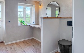 A bathroom with a wooden floor, a window with a plant, a round mirror, and a toilet. - Cozy home in Højer with WiFi (Højer)