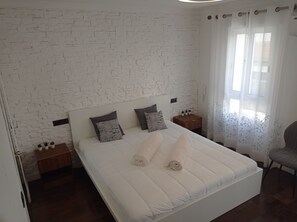 Ein Schlafzimmer mit einer weißen Ziegelwand, einem Bett mit weißer Bettwäsche, zwei Nachttischen mit Lampen und einem Fenster mit durchsichtigen Vorhängen.