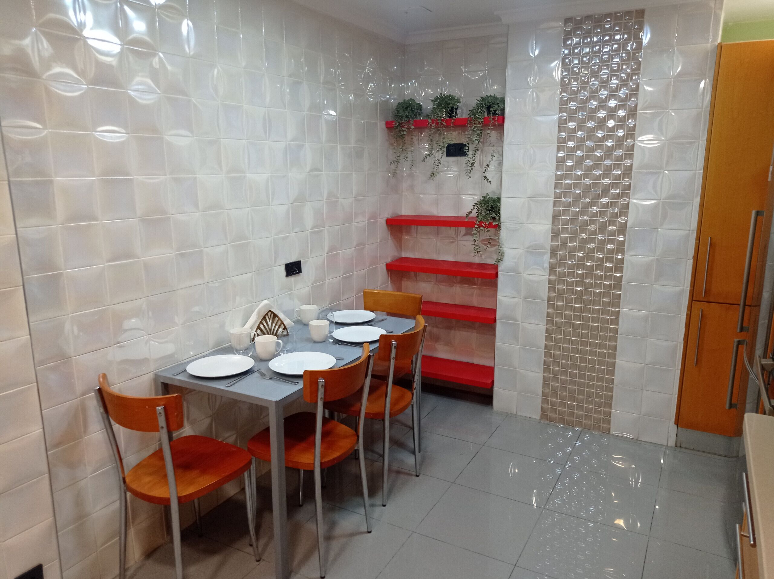 Comedor con una mesa lista para cuatro personas, rodeada de sillas, y un estante rojo con plantas.