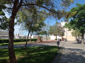 Un parc amb arbres, una zona de jocs i un edifici al fons.