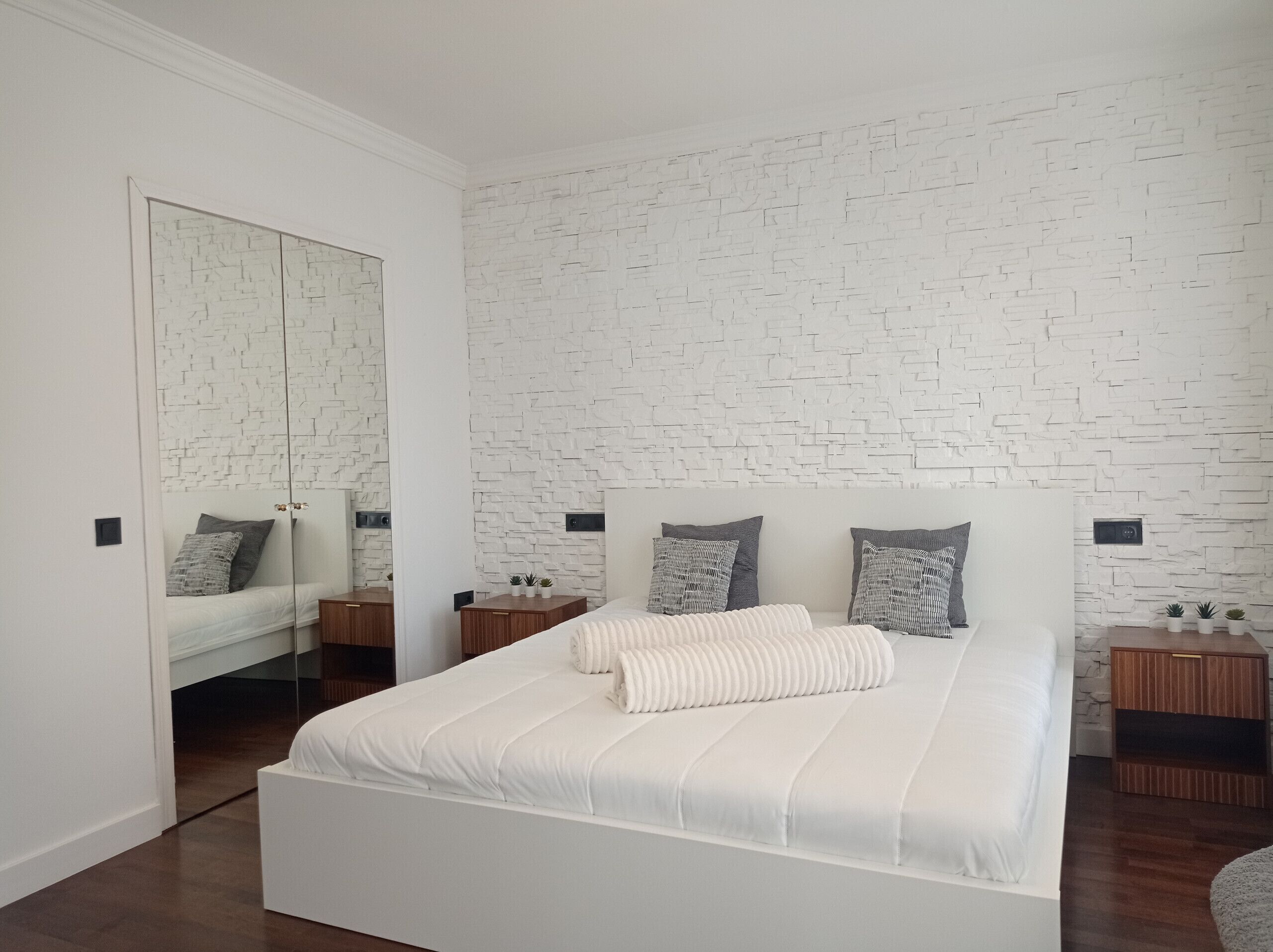 A modern bedroom with a white brick-patterned wall, a large mirror, a bed with white bedding, and two bedside tables with small plants.