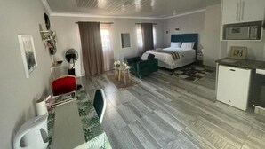 A hotel room with a bed, a desk, a chair, a small table with flowers, a fan, and a kitchenette.