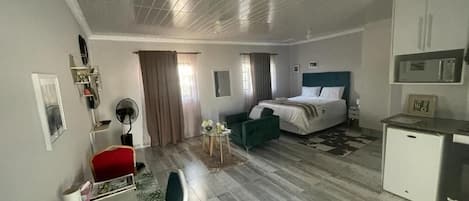 A hotel room with a bed, a desk, a chair, a small table with flowers, a fan, and a kitchenette.