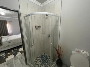 A bathroom with a glass-enclosed shower, a toilet, and a bed in the adjacent room. - OlimG Stays (KuGompo City)
