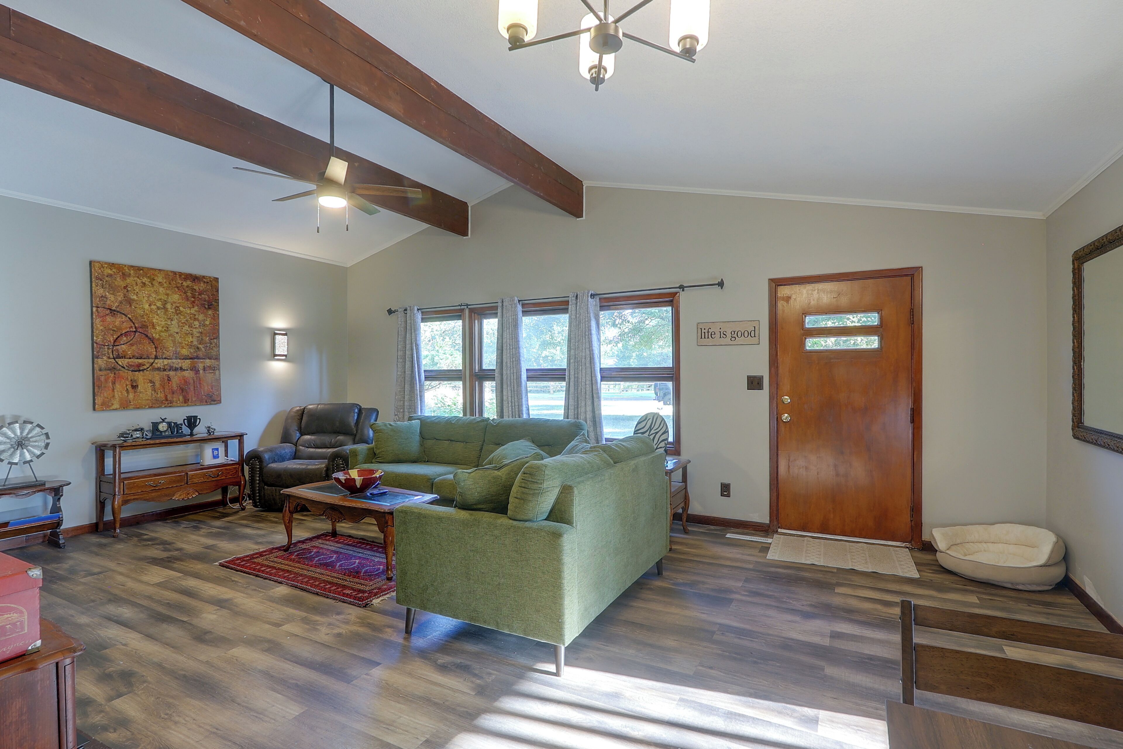 A living room with a green sofa, a leather chair, a wooden coffee table, and a red rug. There is a wooden door with a sign, a window with curtains, and a ceiling fan.
