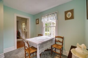 A dining area with a table set for two, two wooden chairs, a lace tablecloth, and a window with a floral valance.