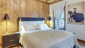 A bedroom with a wooden ceiling, a bed with white bedding, two bedside lamps, and a painting of a woman on a horse.