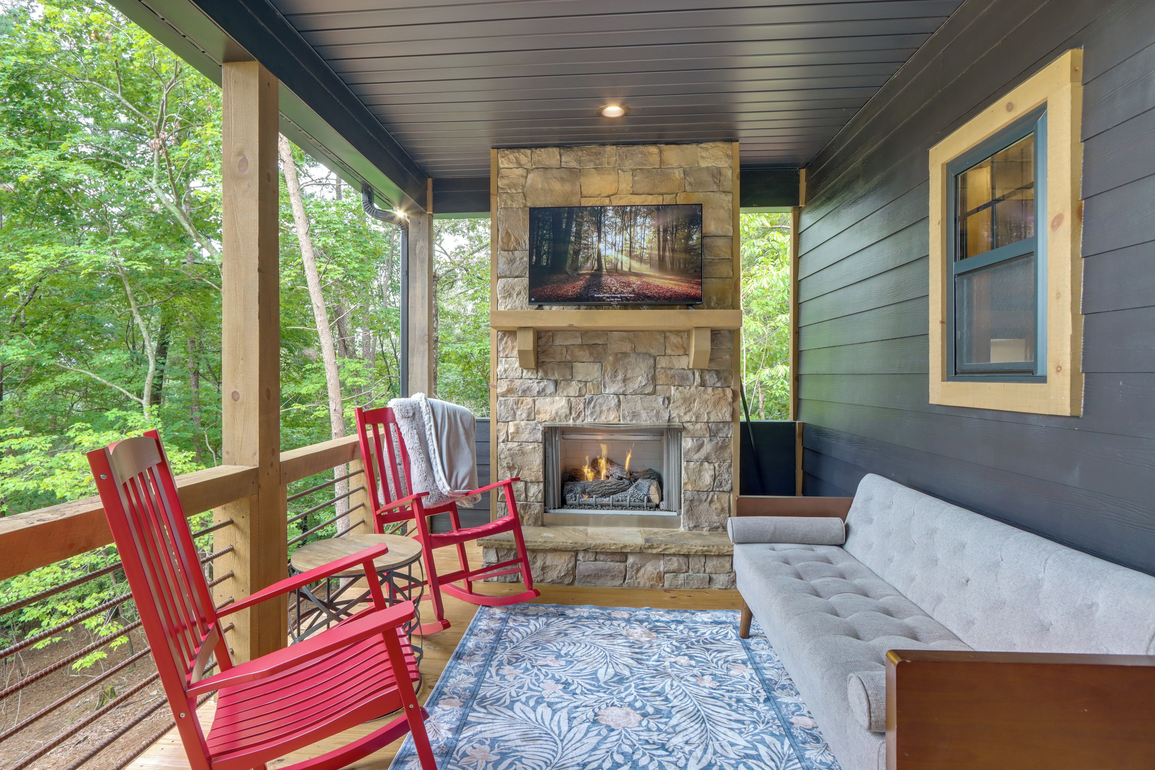 A covered porch with a stone fireplace, red chairs, a grey sofa, and a television mounted on the wall.