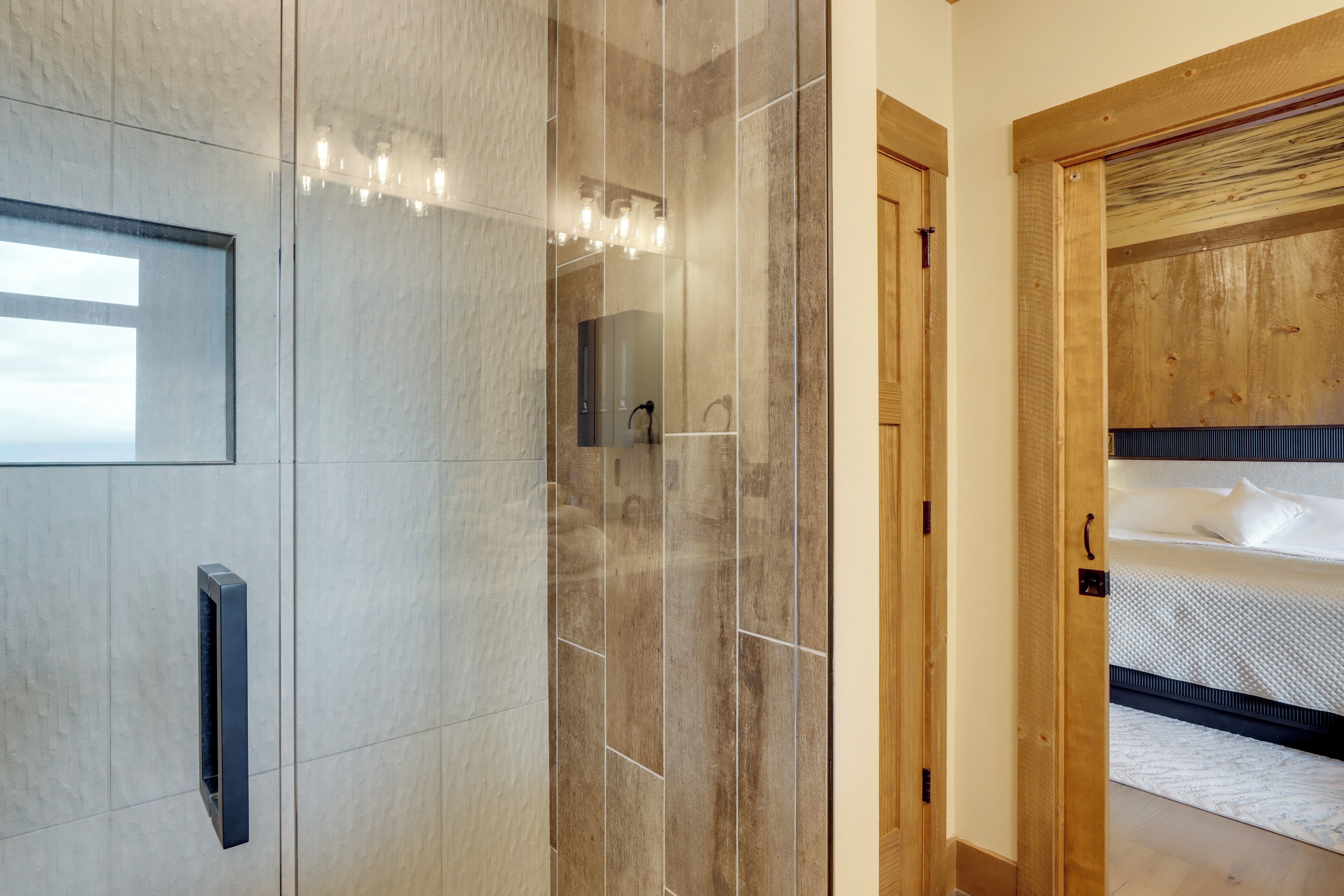 A modern bathroom with a glass shower enclosure, a wooden door leading to a bedroom with a bed and a nightstand.