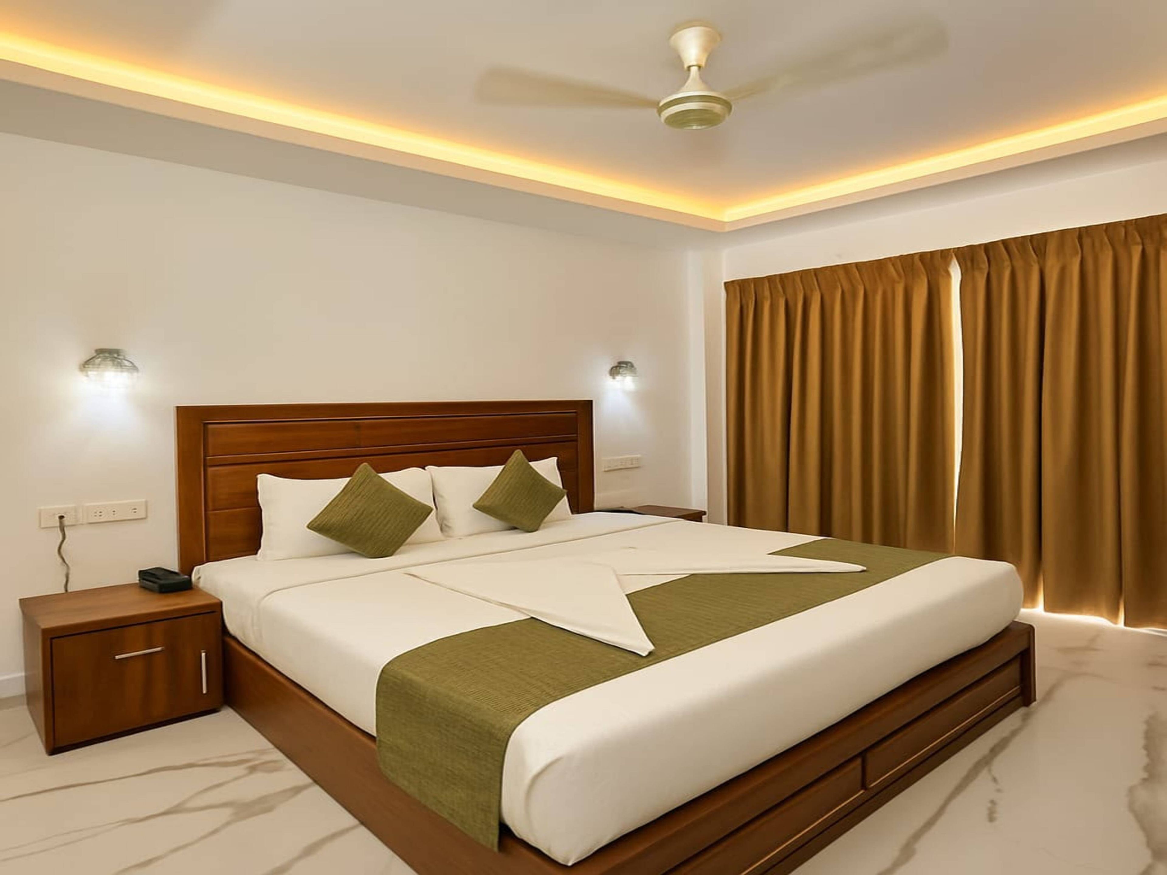 Une chambre d’hôtel équipée d’un lit, d’une table de chevet, d’une lampe fixée au mur, d’un ventilateur de plafond et d’une fenêtre avec des rideaux.