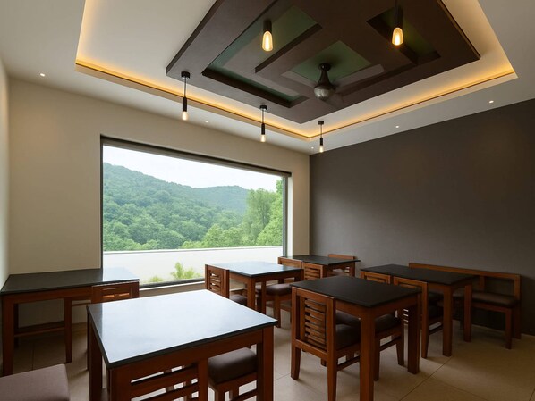 A dining area with multiple tables and chairs, a large window offering a view of a forested landscape, and a modern ceiling design with recessed lighting. - Tripple F Inn - A Luxury Stay in Munnar (Devikolam)
