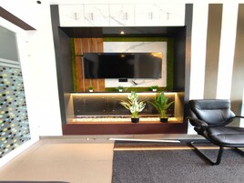 Una zona de espera moderna con un televisor de pantalla plana montado en la pared, un área de descanso con un sillón de cuero negro y un arreglo decorativo de plantas.