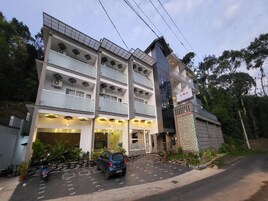 Um hotel de vários andares com design moderno, apresentando varandas em cada pavimento, uma área de estacionamento com alguns carros e uma entrada bem iluminada.