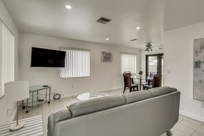 A modern living room with a grey sofa, a flat-screen TV mounted on the wall, and a dining area with a glass table and chairs. - FIU Basecamp: Miami Home w/ Fenced Yard! (Miami)