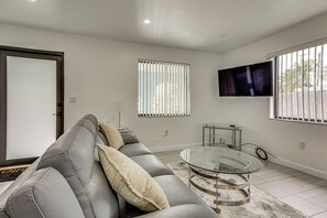 A modern living room with a grey leather sofa, a glass coffee table, and a TV mounted on the wall. - FIU Basecamp: Miami Home w/ Fenced Yard! (Miami)