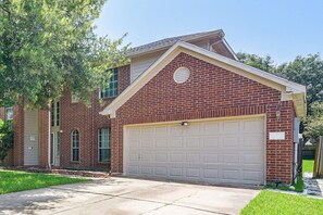 A two-story brick house with a front garage, a small porch, and a well-maintained lawn. - Theater Room & Yard | Near Kemah Boardwalk (Dickinson)
