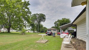A backyard with a lawn, a table, chairs, a covered patio area, and a red vehicle parked in the distance.