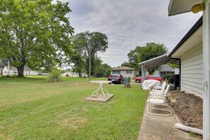 A backyard with a lawn, a table, chairs, a covered patio area, and a red vehicle parked in the distance.
