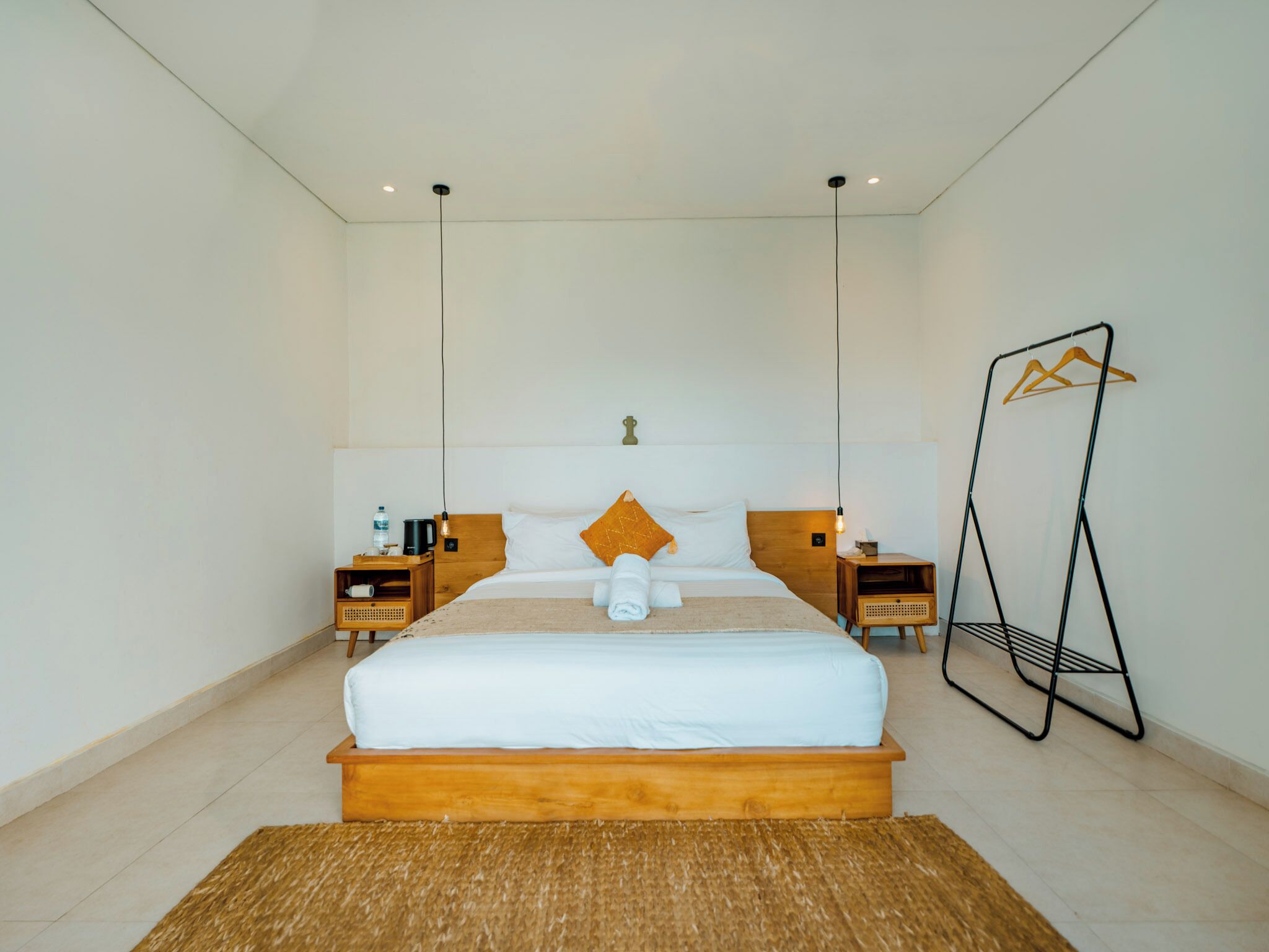 A modern hotel room with a large bed, wooden headboard, and a hanging light fixture. There are two bedside tables with a water bottle and a coffee maker on one, and a small table with a lamp on the other.
