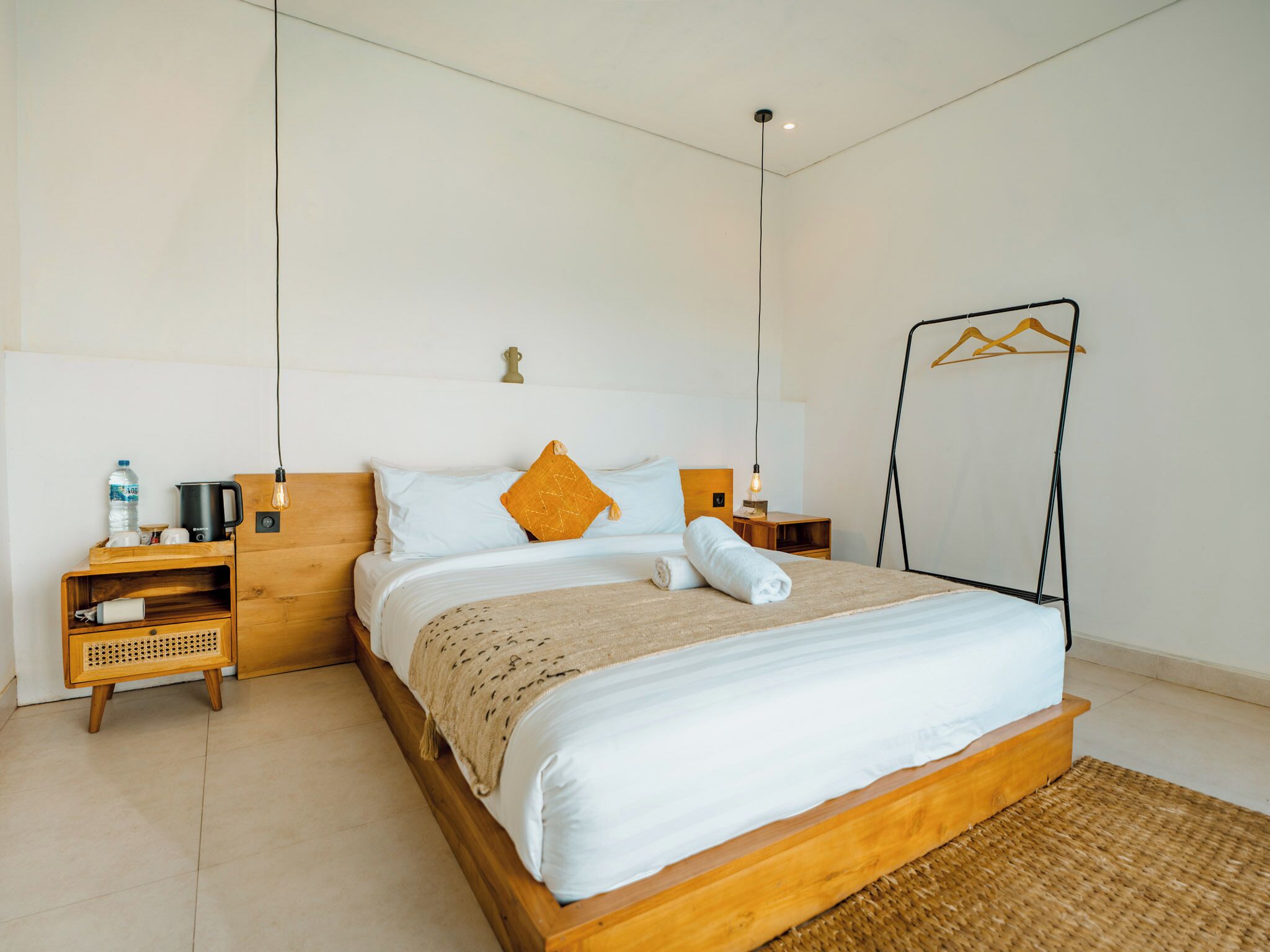 Un dormitorio con cama de madera, ropa de cama blanca, una mesita de noche de madera, una botella de agua, un hervidor y un perchero.