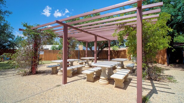 Una pérgola con mesas y bancos de piedra debajo, rodeada de árboles y una valla de madera.