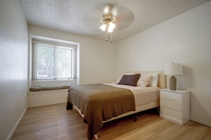 Ein Schlafzimmer mit Bett, Nachttisch, Lampe, einem Fenster mit Jalousien und Deckenventilator.
