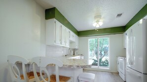 Una cocina con armarios blancos, encimera blanca, un cubo de basura blanco y un refrigerador blanco.