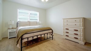 Un dormitorio con cama, mesita de noche, cómoda, ventana con persianas y ventilador de techo.