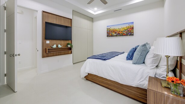 Ein Schlafzimmer mit einem Bett, einem Deckenventilator, einem an der Wand montierten Fernseher, einem Gemälde und einem Nachttisch mit Lampe und Blumen.