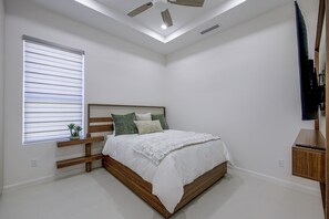 Ein Schlafzimmer mit Bett, einem Fenster mit Jalousien, einem Deckenventilator und einem Fernseher auf einem Holztisch.