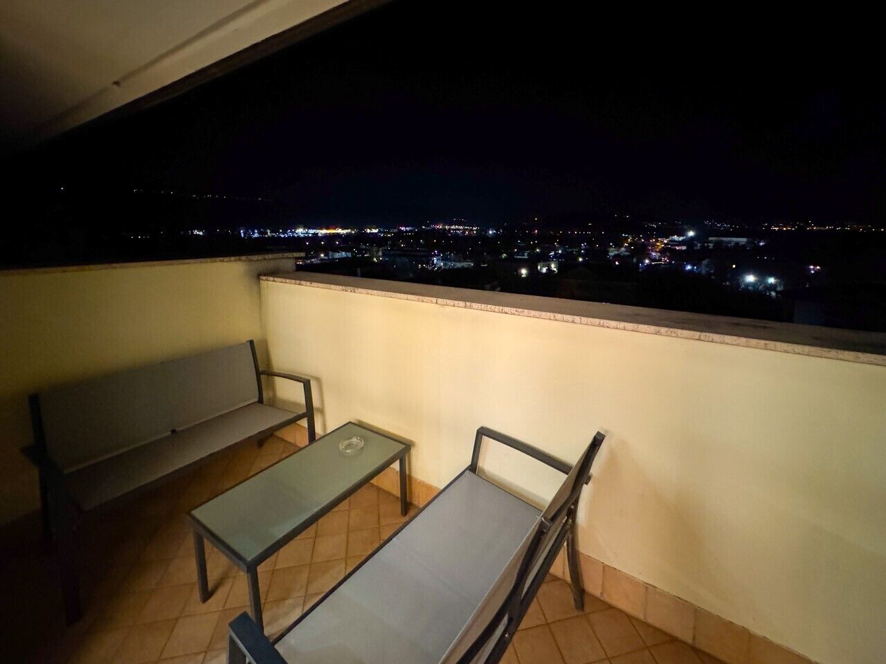 A balcony with a bench and two tables, overlooking a cityscape at night.