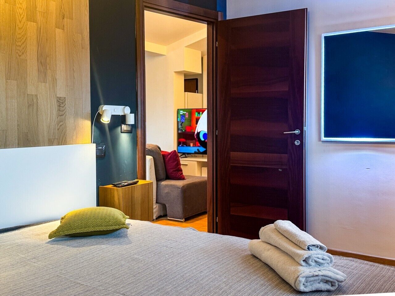 A hotel room with a bed, a bedside table, a lamp, a TV, and a door leading to another room.