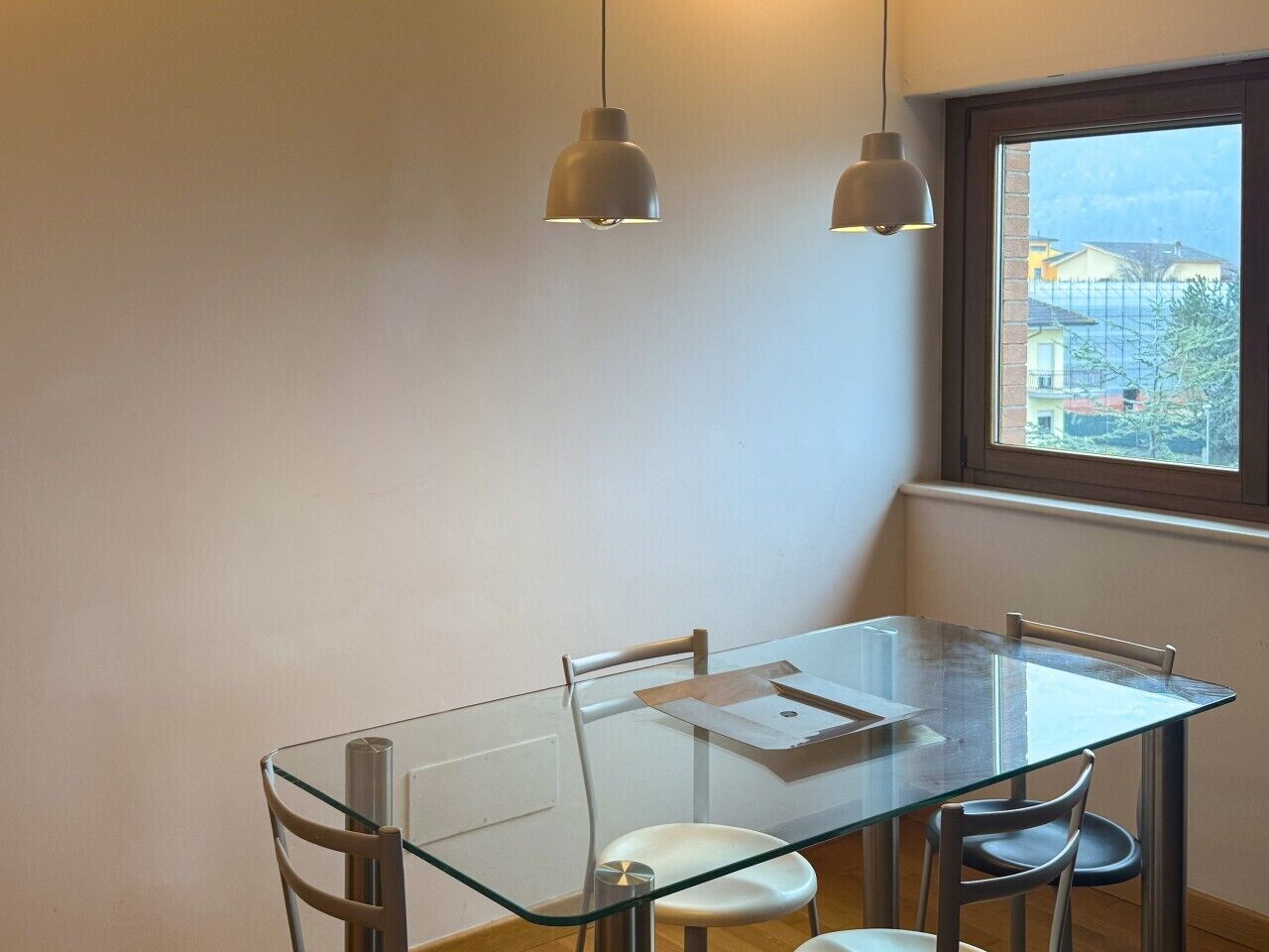 A dining area with a glass table, four chairs, two pendant lights, and a window with a view of buildings.