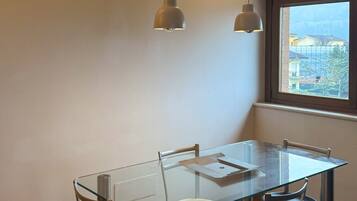 A dining area with a glass table, four chairs, two pendant lights, and a window with a view of buildings.
