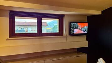 A room with a television mounted on the wall, a window with a view of buildings, and a wooden cabinet below.