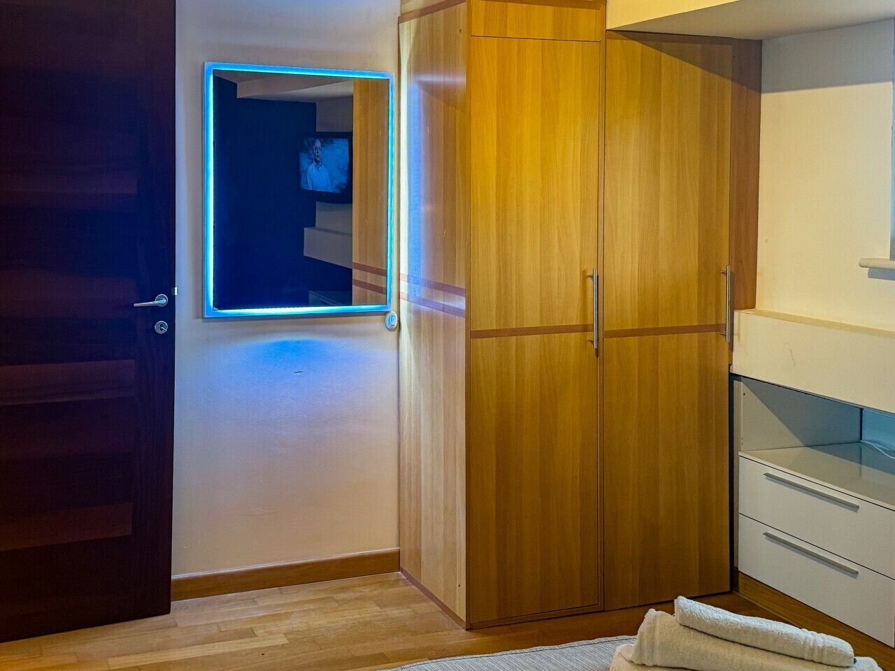 Quarto com guarda-roupa de madeira, espelho com luz neon, armário branco e chão de madeira.