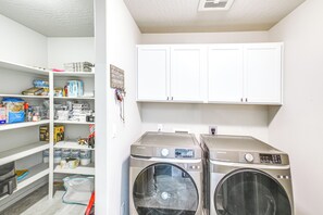 A laundry room with two front-loading washing machines, a shelf with various cleaning supplies, and a welcome sign on the wall. - 3 Mi to Spokane River! Furnished Patio & Yard (Post Falls)