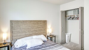 Une chambre à coucher avec une tête de lit en bois, un lit recouvert d’une literie blanche, une table de chevet avec une lampe et une armoire où sont suspendus des vêtements.