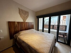 A bedroom with a wooden headboard, a large bed with a textured bedspread, and a sliding glass door leading to an outdoor area. - Condominium in Tulum, Mexico (Tulum)