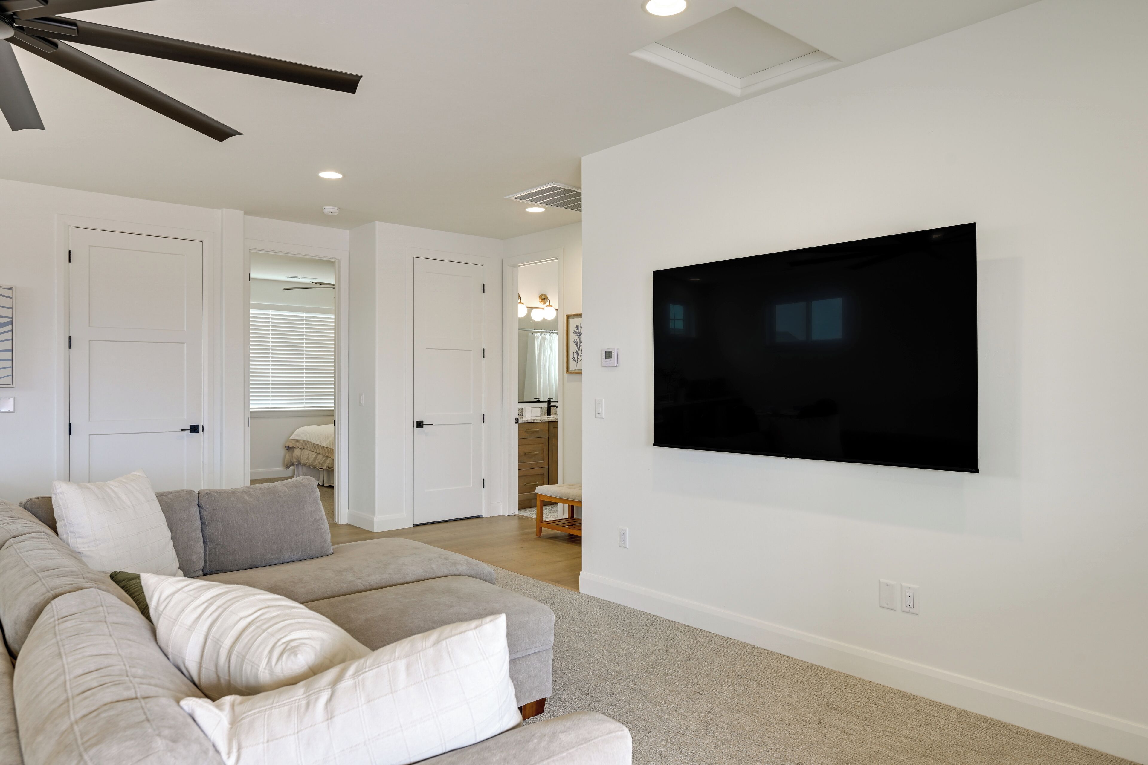 A modern living room with a flat-screen TV mounted on the wall, a ceiling fan, and a large grey sofa with multiple cushions.