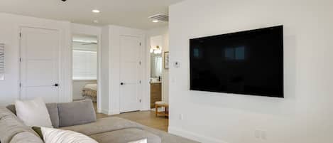 A modern living room with a flat-screen TV mounted on the wall, a ceiling fan, and a large grey sofa with multiple cushions.