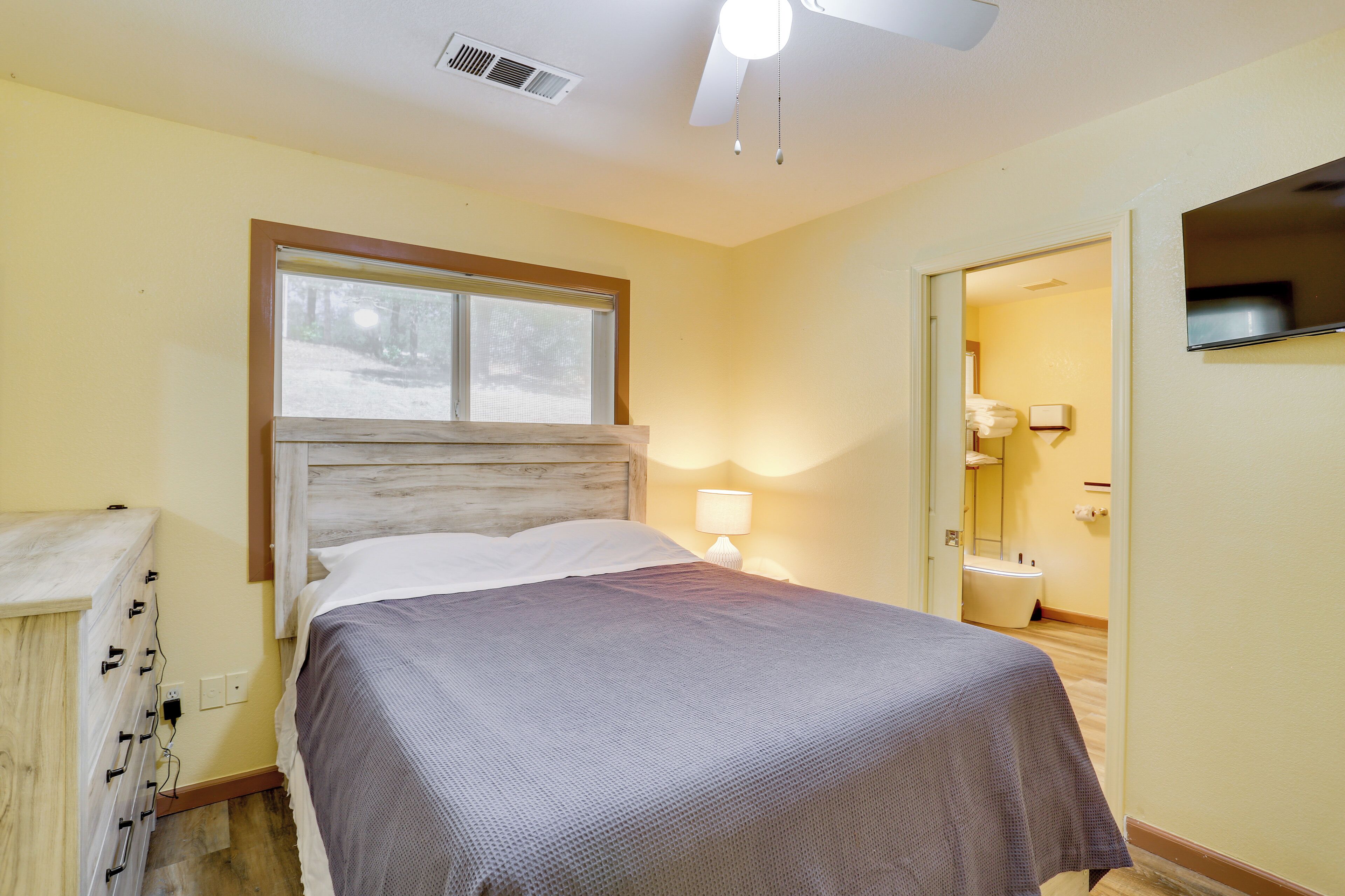 Une chambre d’hôtel avec un lit, une table de chevet avec une lampe, une fenêtre avec des stores, un ventilateur de plafond et une salle de bain visible à travers une porte ouverte.