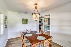 A dining area with a wooden table and chairs, a chandelier, and a kitchen in the background.