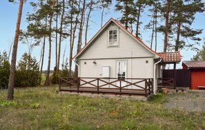 Eine kleine, eingeschossige Holzhütte mit einer Holzterrasse, umgeben von hohen Bäumen und einer Wiese.