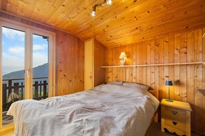 Une pièce en bois avec un lit, une table de chevet, une étagère avec une lampe et une vue sur l’extérieur.
