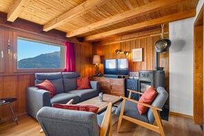 Ein gemütliches Blockhaus-Interieur mit einer Couch, Sesseln, einem Fernseher und einem Kaminofen.