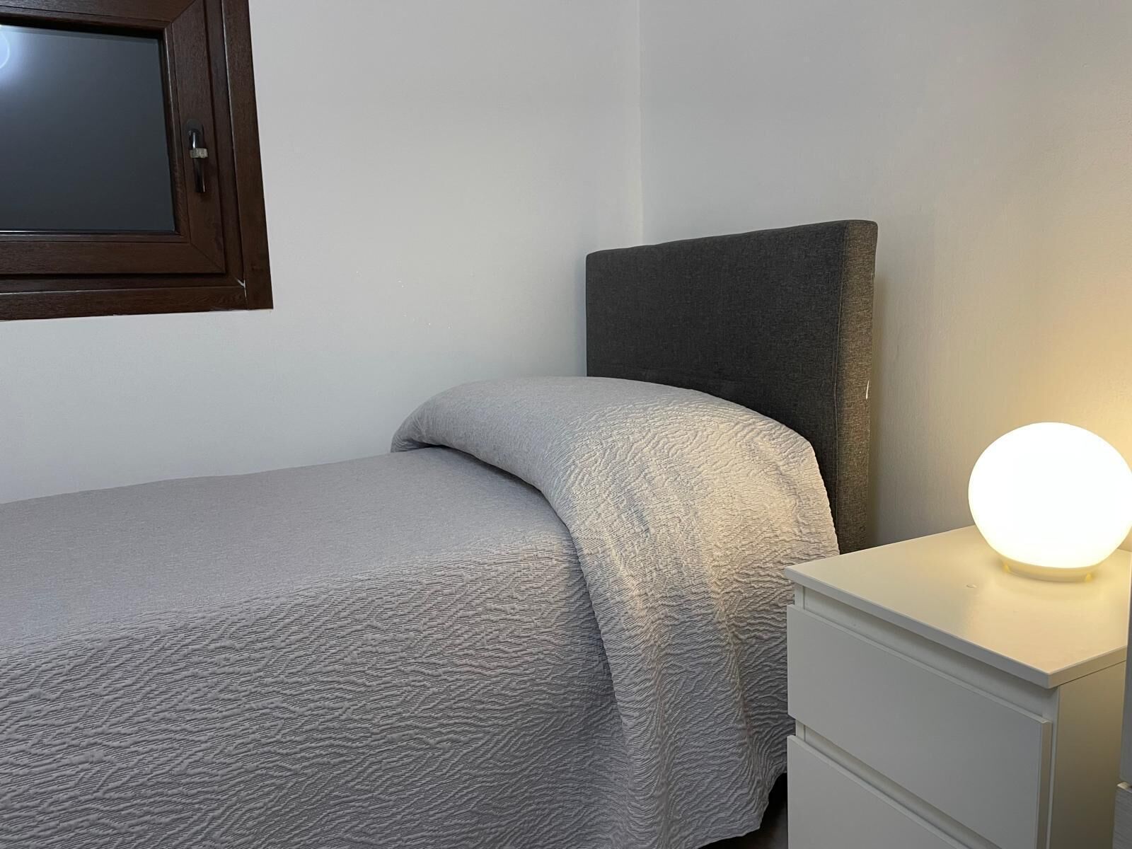 Un lit simple avec une couvre-lit grise à texture, une table de chevet dotée d’une lampe ronde et une tête de lit sombre.