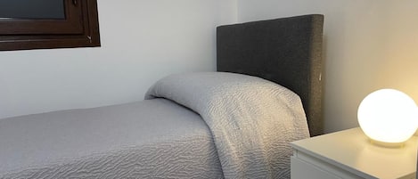 A single bed with a grey textured bedspread, a bedside table with a round light, and a dark headboard.
